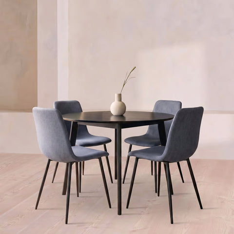 A round black dining table with four gray upholstered chairs and black legs, arranged in a minimalist room with a beige wall and light wood floor. A small white vase with a single stem sits on the table.