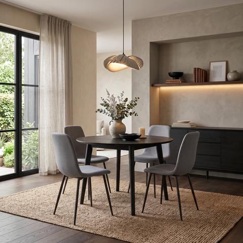 A modern dining room scene featuring a round black table surrounded by four gray upholstered chairs on a woven jute rug, with a pendant light overhead and a built-in shelf in the background.